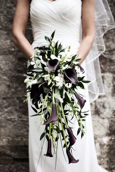 a bride holding a bouquet of flowers and greenery on her wedding day in front of a stone wall