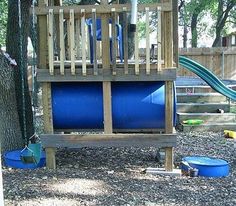 a playground with blue barrels and a slide in the back ground, next to a tree