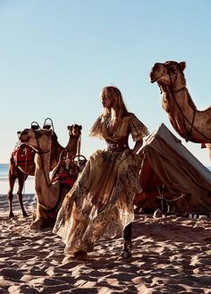 a woman in a long dress standing next to two camels on the sand at the beach