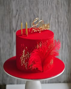 a red cake with gold decorations and feathers on top is sitting on a white pedestal