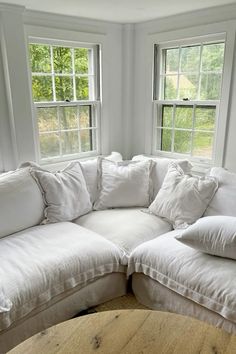 a white couch with pillows on it in front of two windows and a wooden table