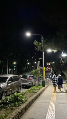 people walking down a sidewalk at night with cars parked on the side walk and street lights in the background