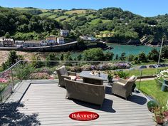 an outdoor deck with wicker furniture overlooking the water and town below it is surrounded by greenery