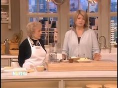 two women standing in front of a kitchen counter with food on the counter and one woman wearing an apron