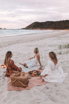 three women sitting on the beach having a picnic with food and drinks in front of them