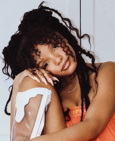 a woman in an orange dress leaning against a white wall with her arms on her chest