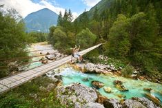 two people standing on a bridge over a river with blue water and green mountains in the background