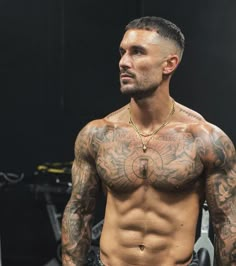 a man with tattoos on his chest standing in front of a gym equipment rack and looking off into the distance