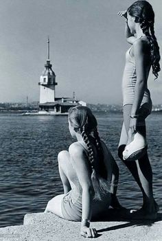 two women in bathing suits sitting on the edge of a body of water with a lighthouse in the background