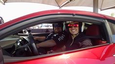 two men sitting in the drivers seat of a red car under an umbrella on a sunny day