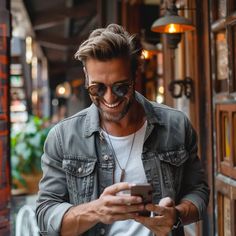 a man wearing sunglasses is looking at his cell phone and smiling while standing in front of a building