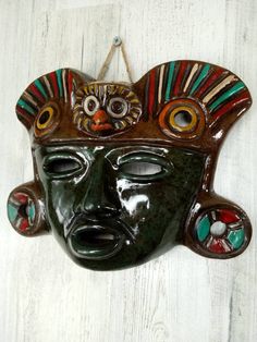 a mask hanging on the wall with eyes and nose painted in green, red, yellow and orange colors