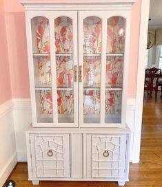 a white china cabinet with flowers painted on the glass doors and gold knobs, in front of a pink wall