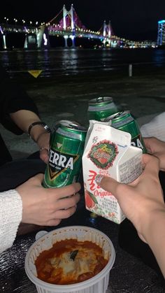 two people holding cans of beer and some food on a table near the water at night