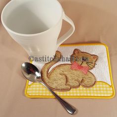 there is a cup and spoon on the table with a cat appliqued on it