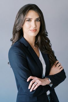 a woman with her arms crossed wearing a black suit and white shirt, standing in front of a gray background