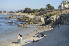 people are standing on the beach next to the water and some houses in the background