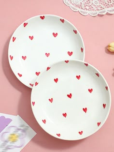 two white plates with red hearts on them next to pink table cloth and doily