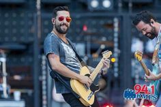 two men are playing guitars on stage at an outdoor music festival, one man is wearing sunglasses and the other has a beard