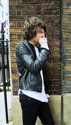 a young man leaning against a brick wall with his hand on his mouth and looking at the camera