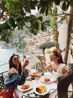 two women sitting at an outdoor table with plates of food in front of the ocean