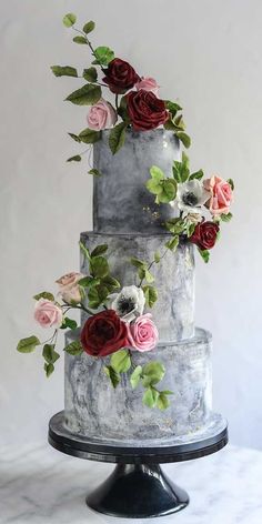 a three tiered cake decorated with flowers and greenery on a black stand against a white background