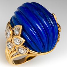 This exquisite cocktail ring is centered with one (1) oval shaped natural lapis lazuli with a carved fluted design. The shoulders of the ring are each accented with six (6) round brilliant cut diamonds bead set into petal shaped setting. The ring measures 23.6mm at the top, rises 15.8mm above the finger, tapering to 2.6mm wide and 1.2mm thick at the base of the shank. It is currently a size 7.25. Luxury Classic Domed Sapphire Ring, Luxury Lapis Lazuli Engagement Rings, Luxury Formal Blue Turquoise Ring, Luxury Gold Sapphire Fusion Ring, Luxury Lapis Lazuli Rings, Elegant Lapis Lazuli Rings For Formal Occasions, Elegant Formal Lapis Lazuli Rings, Elegant Lapis Lazuli Ring With Polished Finish, Carved Stone Jewelry