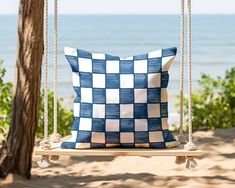 a blue and white checkered pillow sitting on a swing by the beach in front of some trees