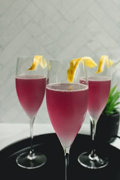 three glasses filled with pink liquid and lemon wedges on a black tray next to a potted plant