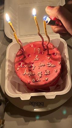 someone lighting candles on a red cake in a white plastic container with the lid open