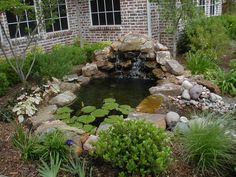 a pond surrounded by plants and rocks in front of a brick building