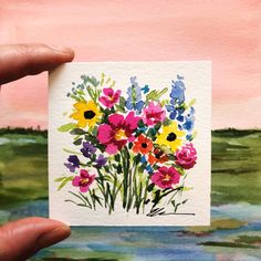 a person holding up a card with watercolor flowers on it in front of a painting