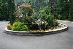 a small waterfall in the middle of a circular garden area with trees and shrubs surrounding it