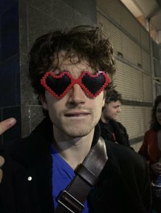 a man with heart shaped glasses on his eyes pointing at the camera while standing in front of a wall