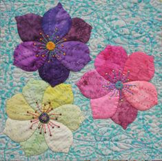 three different colored flowers on a quilted surface