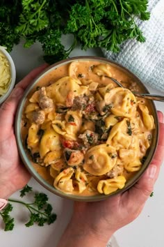 two hands holding a bowl of pasta with meat and spinach on the table next to some parsley