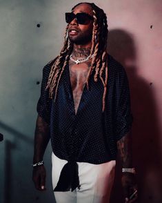 a man with dreadlocks standing in front of a wall wearing sunglasses and a black shirt