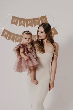 a woman in a white dress holding a baby wearing a pink tulle skirt and standing next to a happy birthday banner