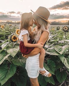 mother and daughter standing in sunflower field at sunset with hat on her head, arms around each other