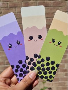 hand holding four different colored bookmarks with faces and eyes on them in front of a brick wall