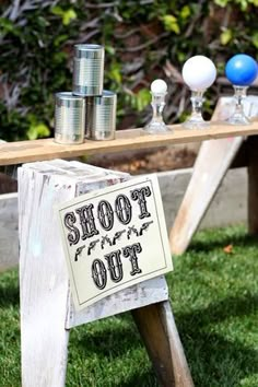 a sign that says shoot out sitting on the grass near an outdoor table with glasses