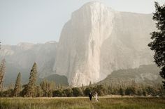 a man and woman are standing in front of a large mountain with trees on it