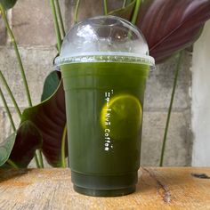 a green smoothie cup sitting on top of a wooden table next to a plant