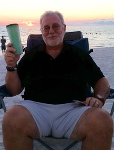 a man sitting in a lawn chair on the beach holding a cup and cell phone