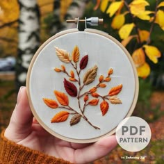 a hand is holding up a embroidery pattern with orange and yellow leaves in the background