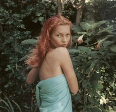 a woman with red hair in a blue dress standing next to trees and bushes, looking at the camera