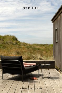 A black outdoor lounge chair with cushion and a black metal outdoor tray table set on a wooden deck patio near grassy dunes