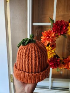 a hand holding up a knitted pumpkin hat in front of a door with flowers