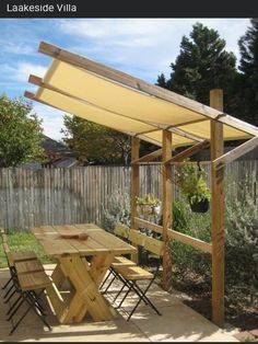 a wooden table sitting under a yellow awning next to a garden area with chairs