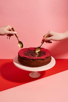 two hands holding spoons over a chocolate cake on a red and pink tablecloth